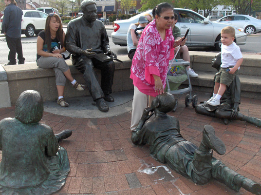 4 photos show life-sized statues on a plaza, one is of Haley sitting on a wall reading a book, there are 3 other statures of children listening to him.  One child statue lies on his stomach looking up at Haley, two others are sitting down.  Standing on Haley's lap and on one of the child statues is a real child.